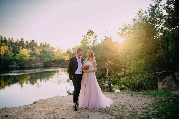 Heureux jeunes mariés debout sur la rive du lac au coucher du soleil — Photo