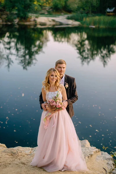 Nowożeńcy, stojąc na brzegu jeziora o zachodzie słońca — Zdjęcie stockowe