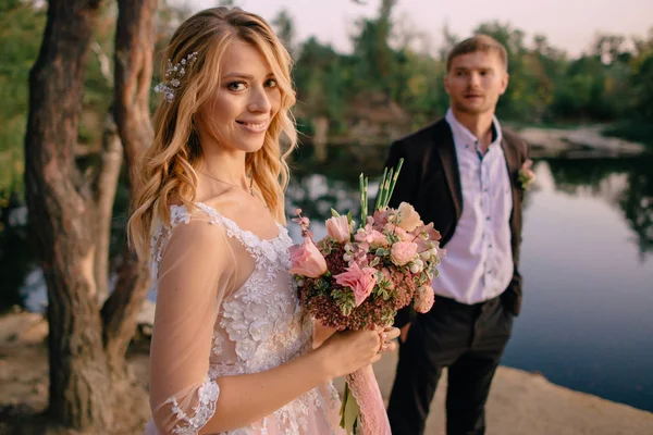 Novomanželé stojí na břehu jezera při západu slunce — Stock fotografie