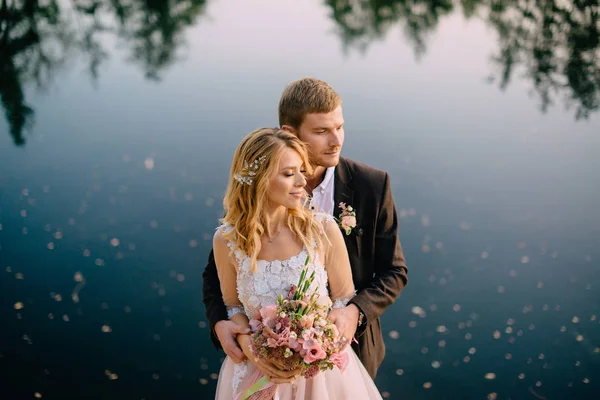 Novomanželé stojí na břehu jezera při západu slunce — Stock fotografie