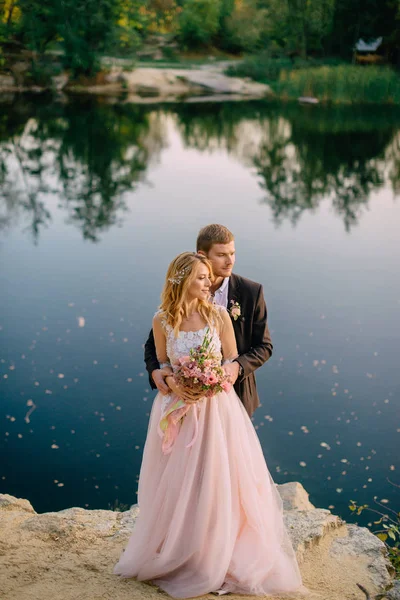 Jeunes mariés debout sur la rive du lac au coucher du soleil — Photo