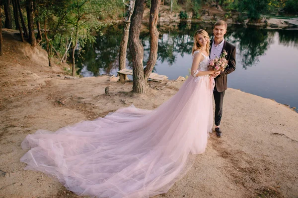 Recém-casados em pé na margem do lago ao pôr do sol — Fotografia de Stock