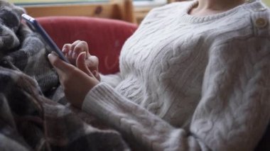 Pencerenin yanındaki rahat sandalyede akıllı telefon kullanan kadınların yakın plan fotoğrafları.