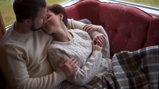Молодий чоловік обіймає дружину, сидячи на дивані вдома — стокове відео