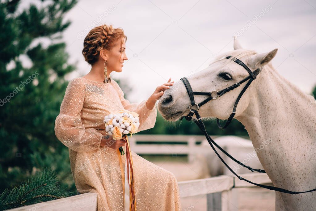 Young bride in a gold dress stroking the head of a white horse