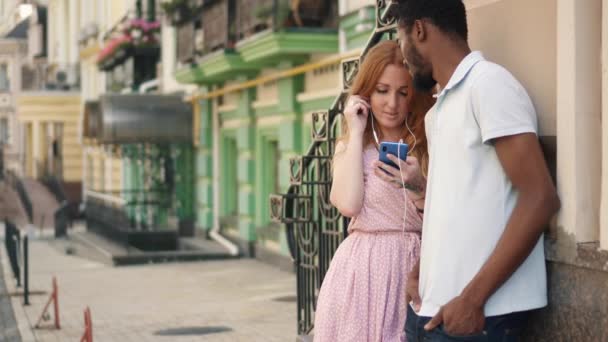 A young woman gives one earphone to her boyfriend to listen to a song together — Stock Video