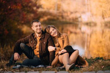 Happy young couple sitting on a lake in autumn park clipart
