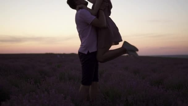 Silhouette Egy fiatalember a karjaiban neveli a barátnőjét, amint egy virágzó levendulamezőn áll napnyugtakor. — Stock videók
