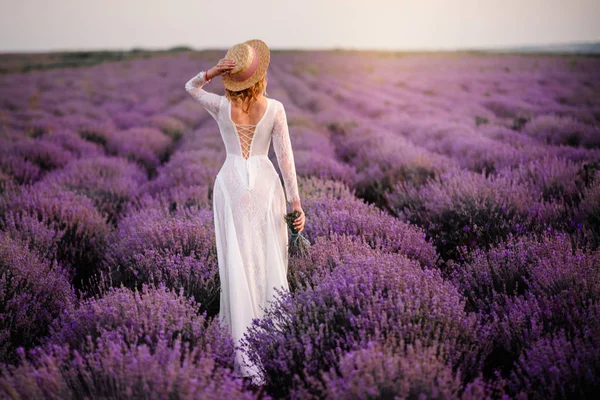 Beyaz elbiseli genç bir kadın gün batımında çiçek açan lavanta tarlasında yürüyor.