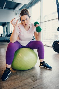 Spor salonunda spor yaparken halterlerle egzersiz yapmaktan yorgun düşmüş şişman bir kadın.