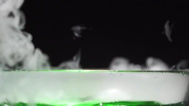 Siyah arka planda cam bir kasede duman çıkaran yeşil sıvı kaynıyor. Detaylı bir yakın plan..