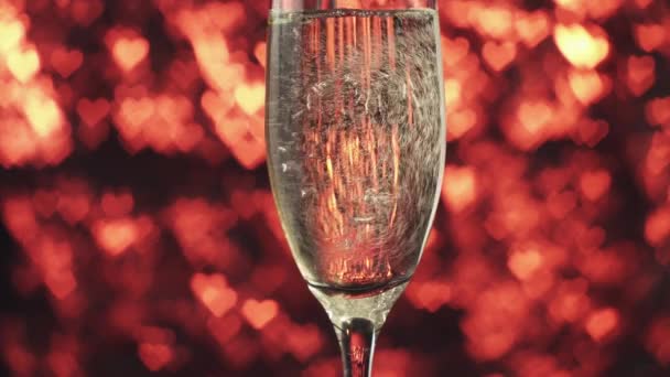 Champagne viene versato in un bicchiere su uno sfondo rosso con molti cuori, da vicino . — Video Stock