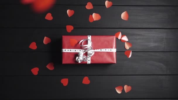 Muchos corazones rojos caen sobre la mesa, en la que yace una caja con un regalo . — Vídeo de stock