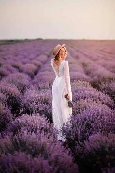 Fehér ruhás fiatal nő sétál a virágzó levendulamezőn naplementekor. — Stock Fotó