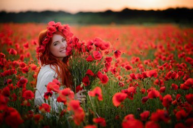 Gün batımında çiçekli tarlada oturan kırmızı haşhaş buketiyle genç ve güzel bir kadın.