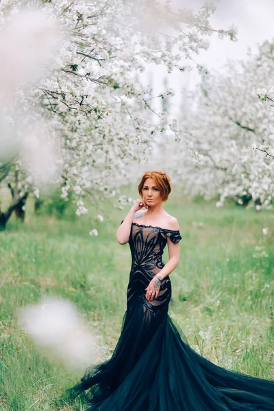 Siyah elbiseli genç bir kadın çiçek açan bir bahçede duruyor.. — Stok fotoğraf