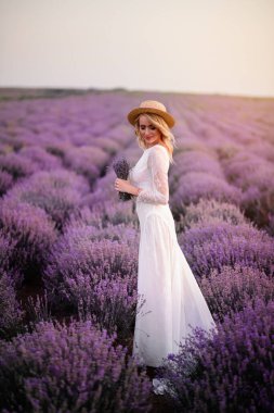 Beyaz elbiseli genç bir kadın gün batımında çiçek açan lavanta tarlasında yürüyor.