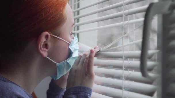 En kvinna i skyddsmask tittar ut genom fönstret.. — Stockvideo