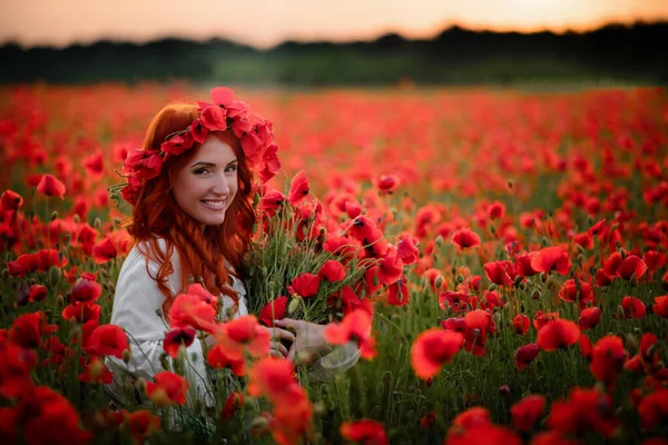 Jovem bela fêmea com buquê de papoilas vermelhas sentadas no campo florido ao pôr do sol — Fotografia de Stock