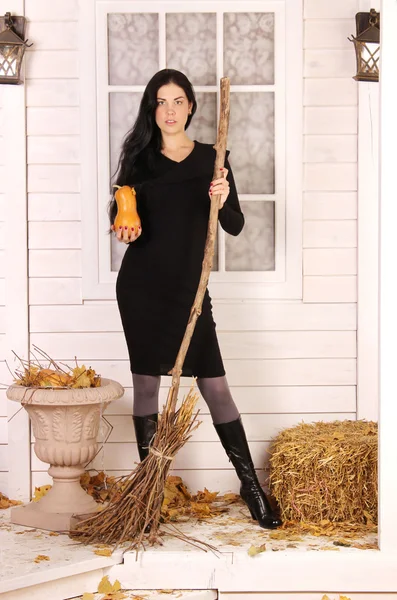 Femme en robe noire tenant une citrouille et un balai sur fond de maison en bois — Photo