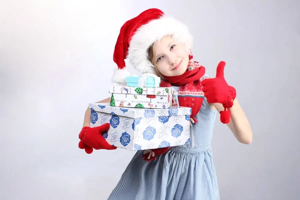 Menina em Santa chapéu ajudante gesticulando polegares para cima e segurando presentes de Natal — Fotografia de Stock