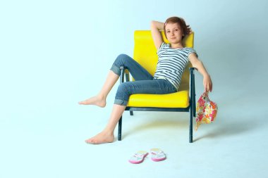 Sevimli genç kız, mavi kot pantolon ve sarı kol-sandalye üzerinde mavi arka plan üzerinde oturan çizgili tişört giyiyor.