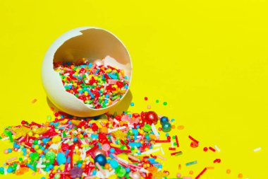 Sarı arka planda kırılan beyaz yumurta kabuğunda çok renkli küçük tatlılar. Paskalya, tatiller, yemek konsepti. Çekimser renkli gıda arka planı.