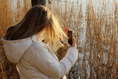 Beyaz ceketli genç bir kız dışarıda, arka manzaralı bir fotoğraf çekiyor. Peole, seyahat, doğa konsepti. Instagram için fotoğraf çeken genç bir kız..