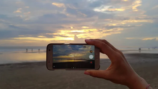 Mano femenina despega en su teléfono inteligente puesta de sol en la playa Fotos De Stock Sin Royalties Gratis