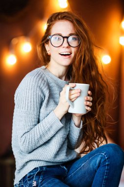 Noel ile odada oturup çay bardağı tutan uzun kızıl saçlı genç gülümseyen kız arka plan üzerinde ışıklar