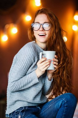 Noel ile odada oturup çay bardağı tutan uzun kızıl saçlı genç gülümseyen kız arka plan üzerinde ışıklar