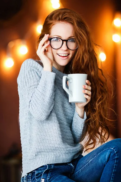 クリスマスの部屋に座って紅茶カップを保持している長い赤い髪を持つ少女笑顔ライトの背景に — ストック写真