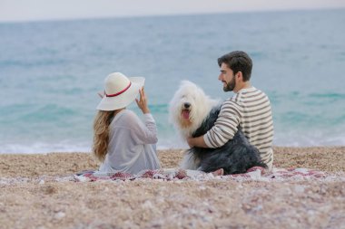Hasır şapka ve sakallı adam ekose deniz kıyısında oturan tüylü köpekle oynayan genç kadın