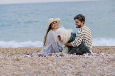 Hasır şapka ve sakallı adam ekose deniz kıyısında oturan tüylü köpekle oynayan genç kadın