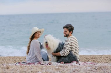 Hasır şapka ve sakallı adam ekose deniz kıyısında oturan tüylü köpekle oynayan genç kadın