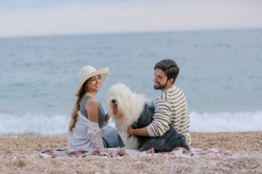 Hasır şapka ve sakallı adam ekose deniz kıyısında oturan tüylü köpekle oynayan genç kadın