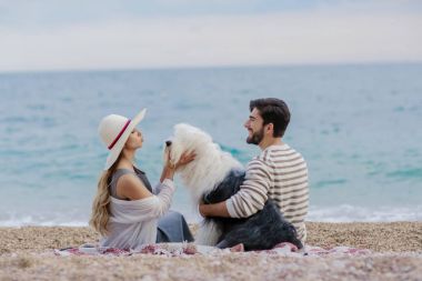 Hasır şapka ve sakallı adam ekose deniz kıyısında oturan tüylü köpekle oynayan genç kadın