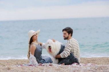 Hasır şapka ve sakallı adam ekose deniz kıyısında oturan tüylü köpekle oynayan genç kadın