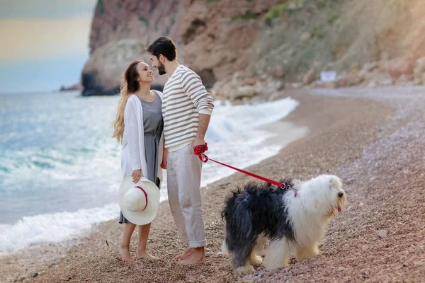 若いカップルがビーチでふわふわの犬を連れて歩いてハグの全長 — ストック写真