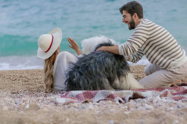 麦わら帽子と髭の男が海岸に格子縞の上に座ってふわふわ犬と遊んでの若い女性 — ストック写真