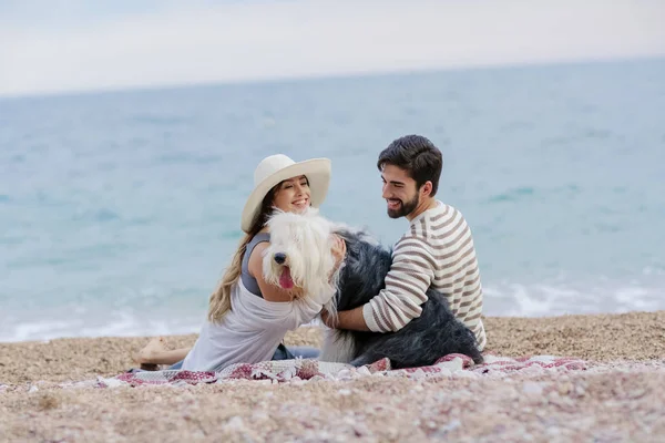 麦わら帽子と髭の男が海岸に格子縞の上に座ってふわふわ犬と遊んでの若い女性 — ストック写真