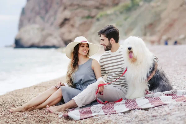 ビーチとお互いに笑みを浮かべて見る格子縞の上に座って犬と休憩の愛の若いカップル — ストック写真