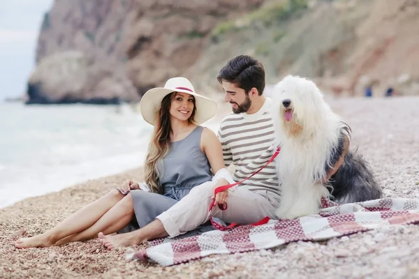 ビーチとお互いに笑みを浮かべて見る格子縞の上に座って犬と休憩の愛の若いカップル — ストック写真