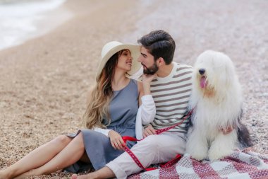 Genç çift ekose plaj ve gülümseyen birbirine bakarak otururken tüylü köpek ile dinlenme aşık
