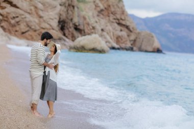 şık genç çift aşık ayakta kumsalda yalınayak cliff ve arka plan, tam uzunlukta dağda ile sarılma