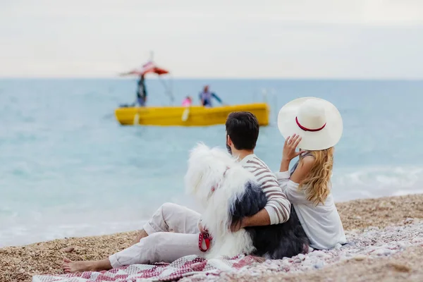 young beard man and beautiful woman in white straw hat resting with fluffy dog on beach with yellow boat swimming in sea on background