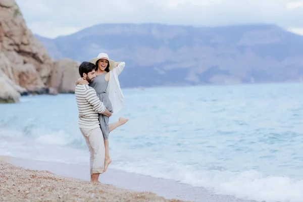 背景には 完全な長さの崖のビーチに立っている彼の美しい女性を持ち上げるを受け入れ幸せな男 — ストック写真