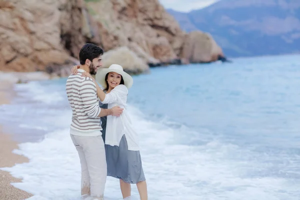 背景の崖のビーチに立っている彼の美しい女性を抱きしめる幸せな男 — ストック写真