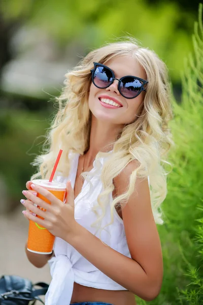 美丽的金发女郎戴着墨镜坐在绿色公园里喝着果汁 — 图库照片