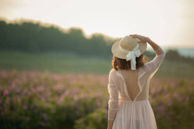 woman in long dress and straw hat standing on green nature background, back view clipart